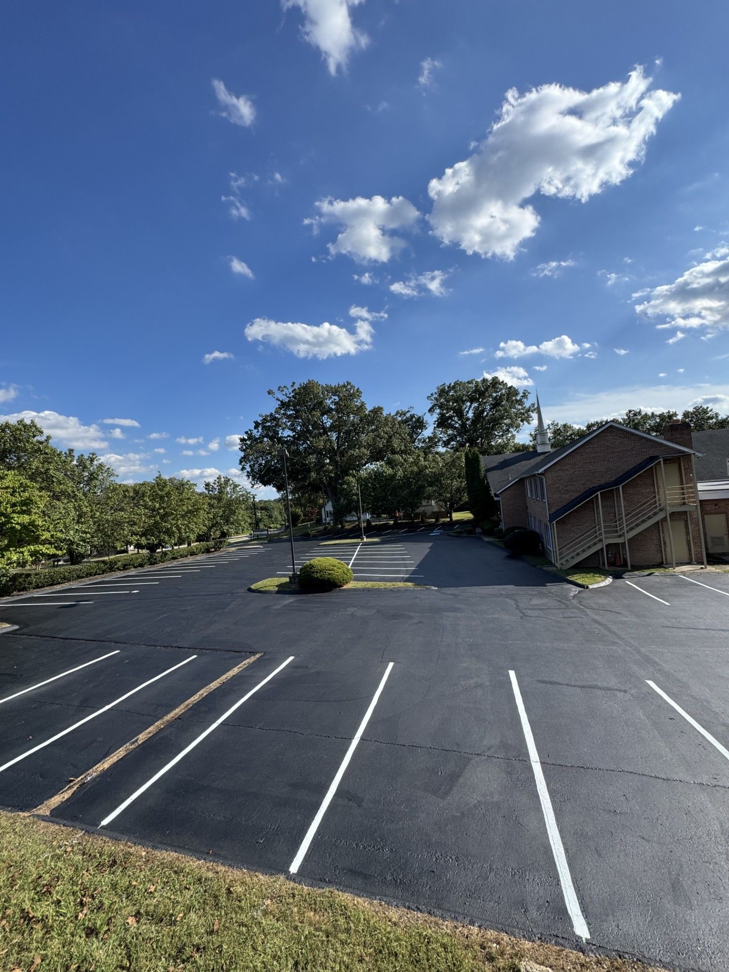 Empty asphalt parking lot on a sunny day, with a blue sky, trees, and a brick building.