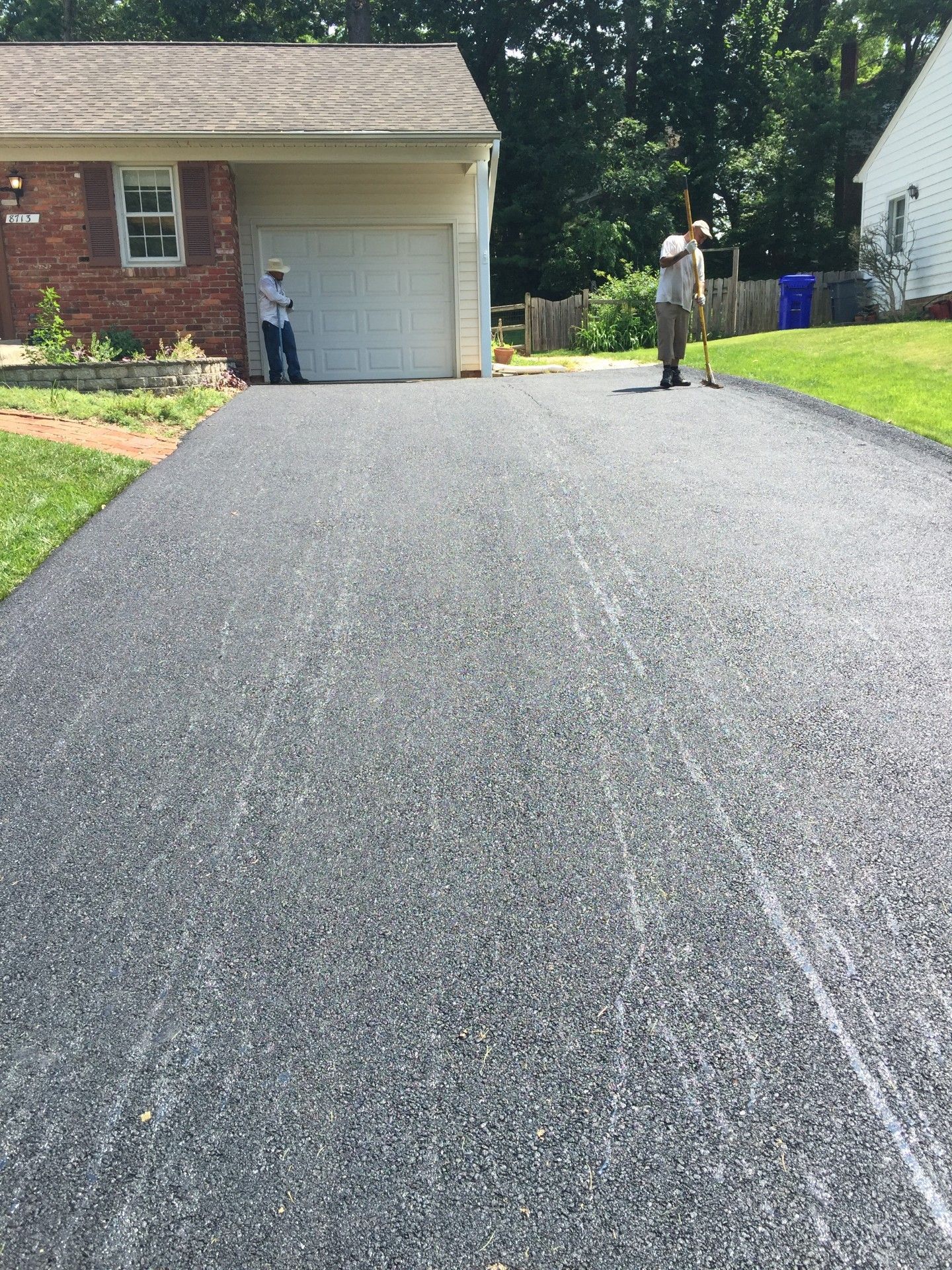Newly paved asphalt driveway; two workers with tools near garage, brick house in background.
