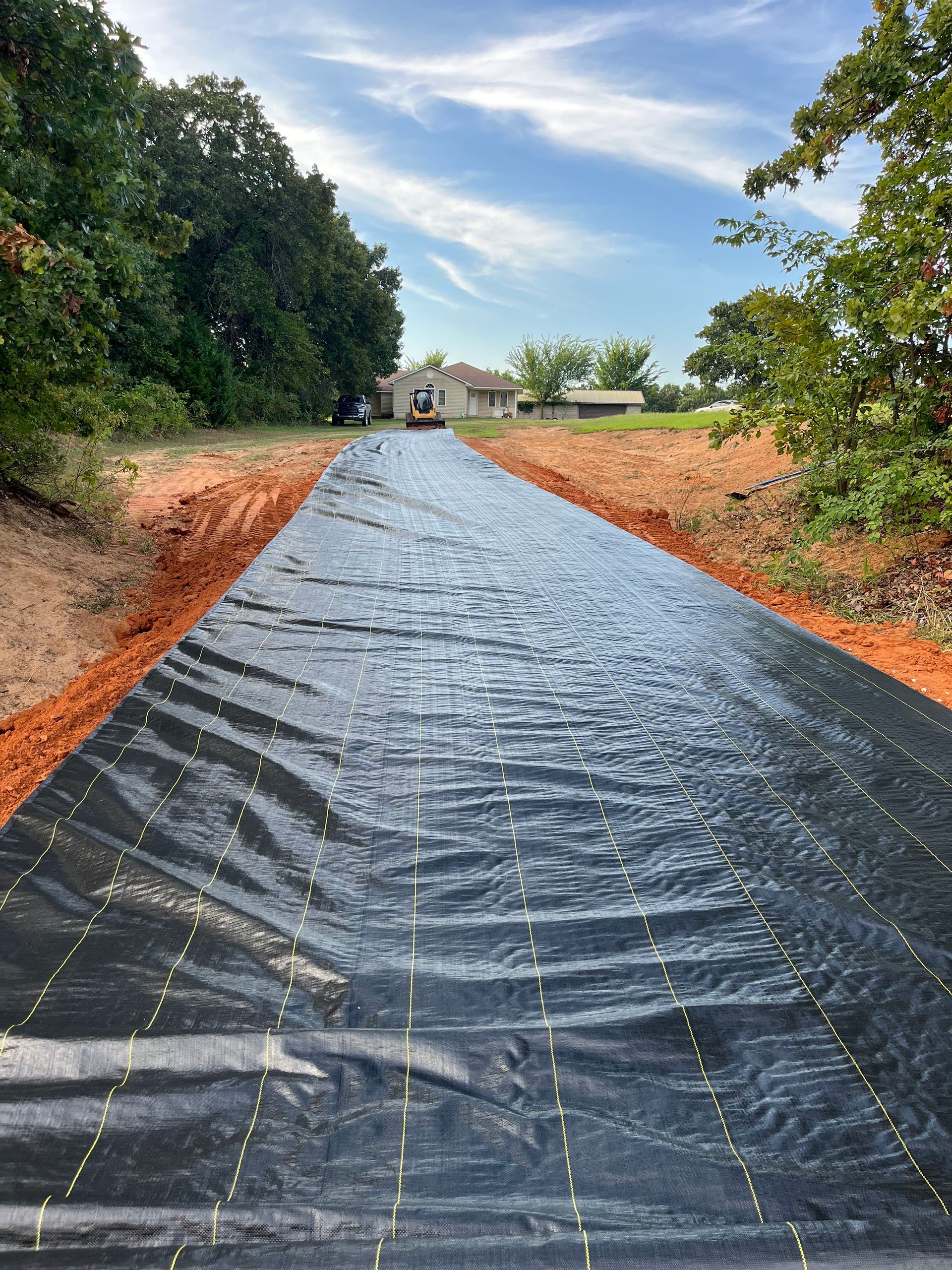 Black landscape fabric laid out on a dirt path, leading towards a distant house and trees under a blue sky.