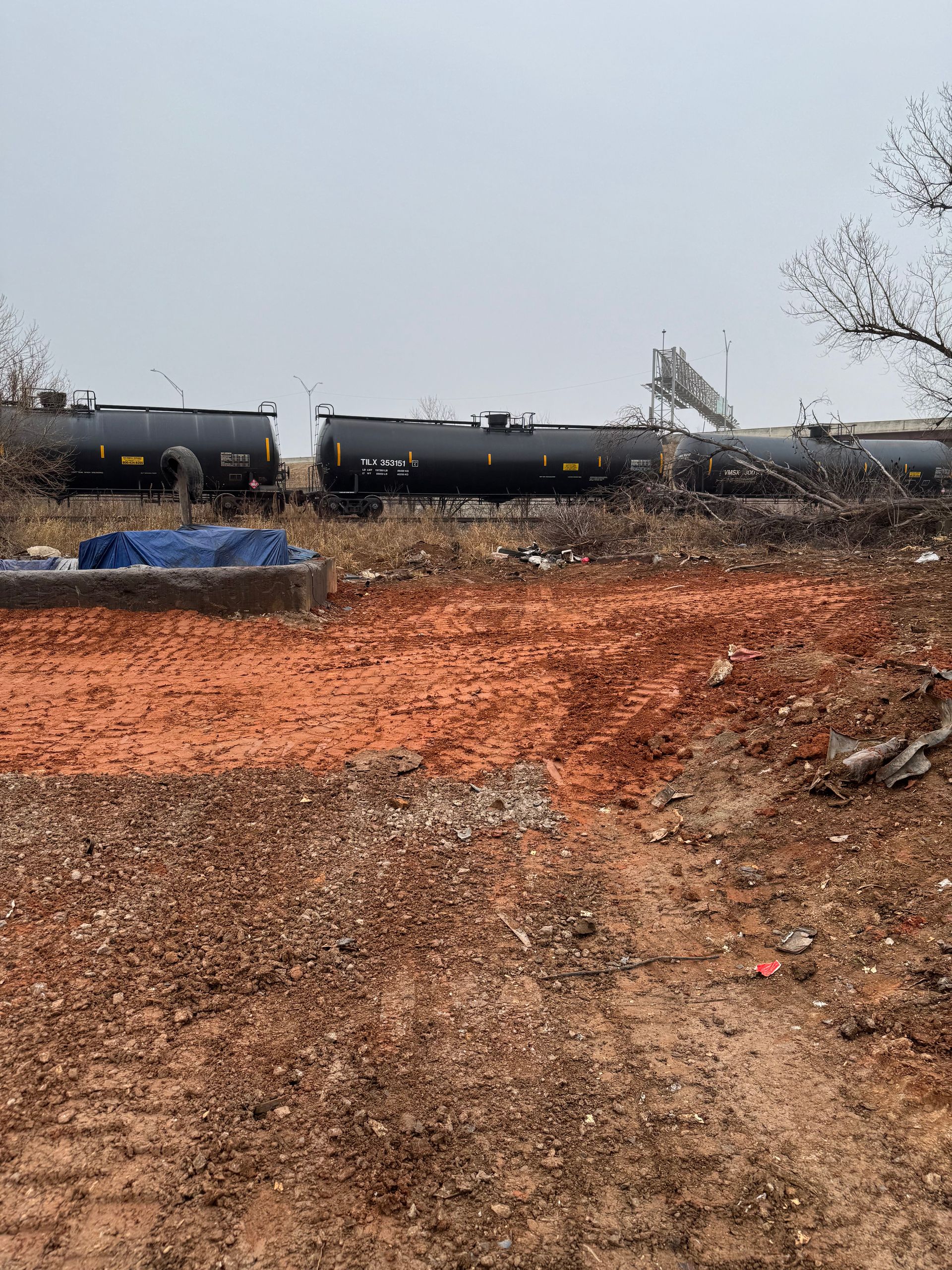Railroad tank cars sit near reddish-brown dirt. A person works near the cars under an overcast sky.