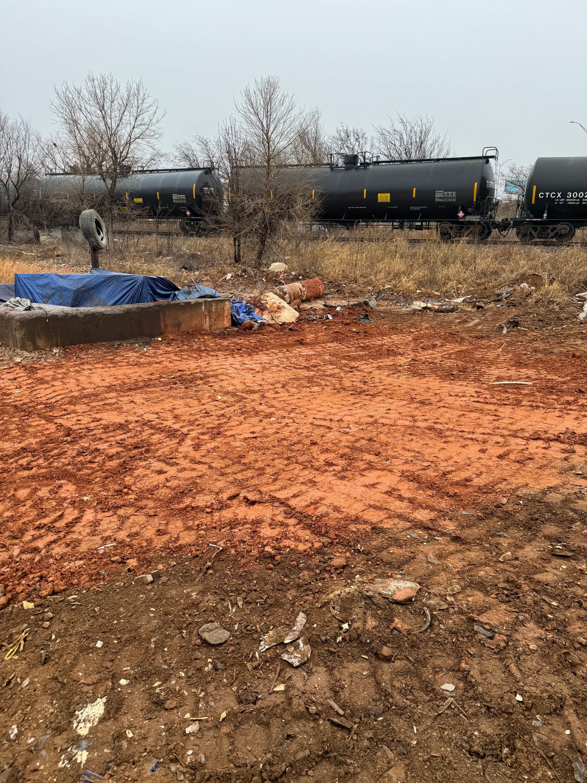 An open area of red-brown material, a person near a blue tarp, and tanker cars in the background on an overcast day.