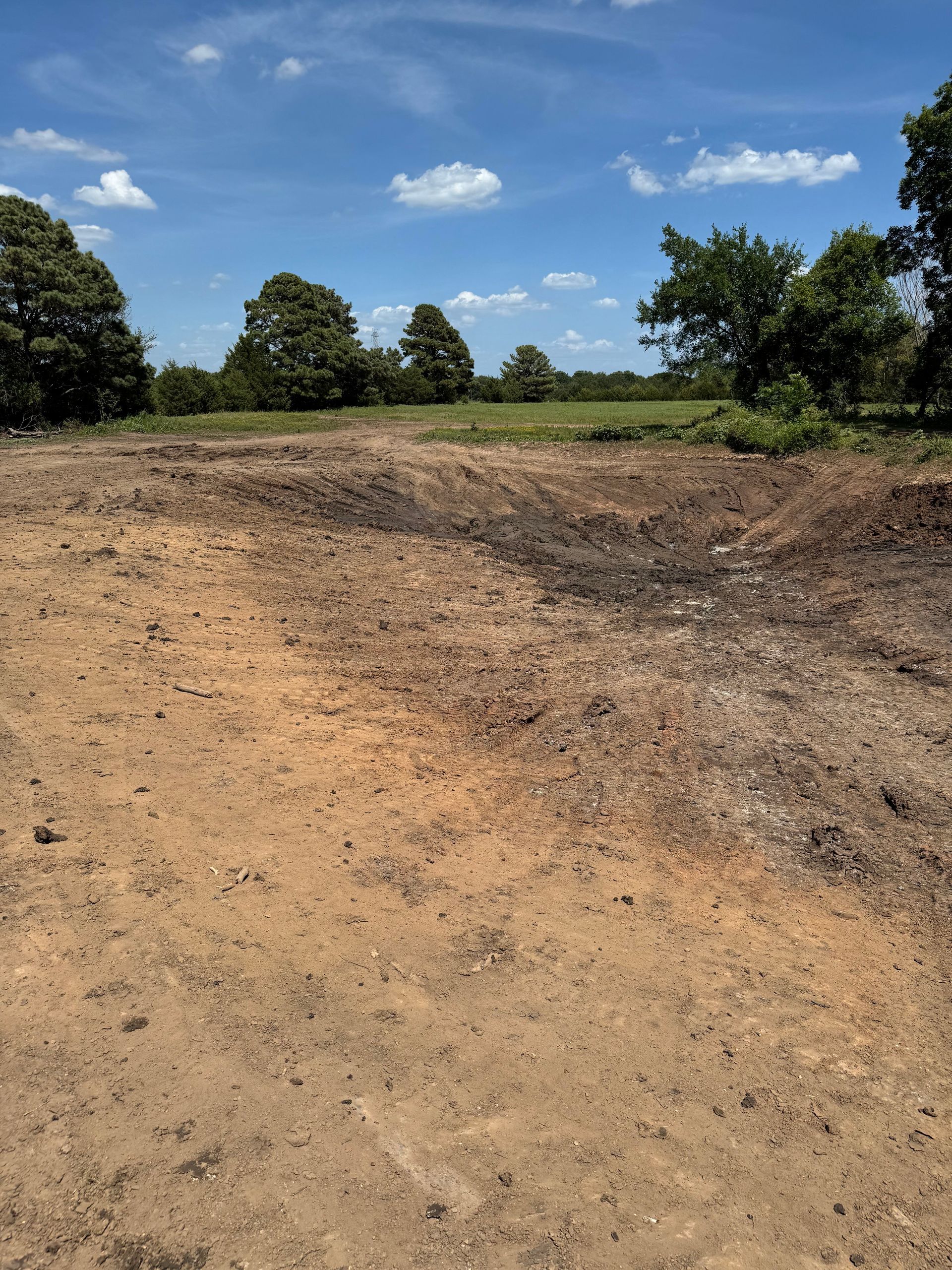 Open dirt lot with trees in the background under a blue sky with scattered clouds.