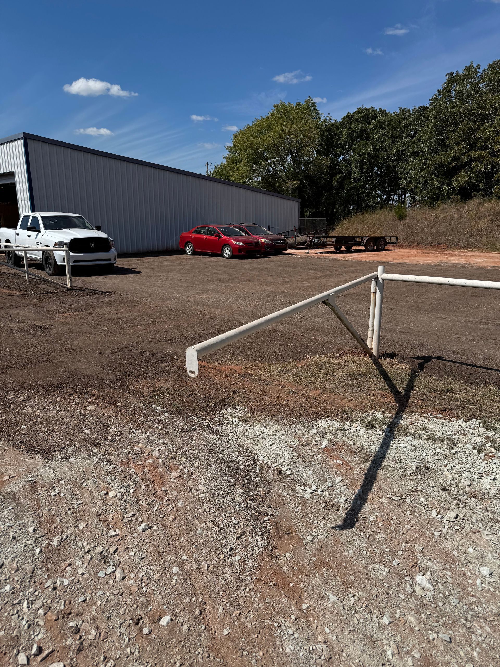 A red car and a white truck parked near a metal building on a gravel lot under a blue sky.