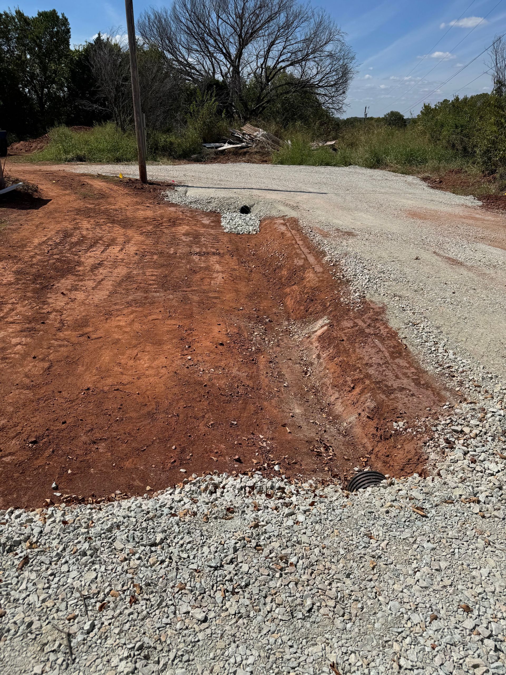 Gravel path merging with red dirt, a drain, and vegetation under a blue sky.