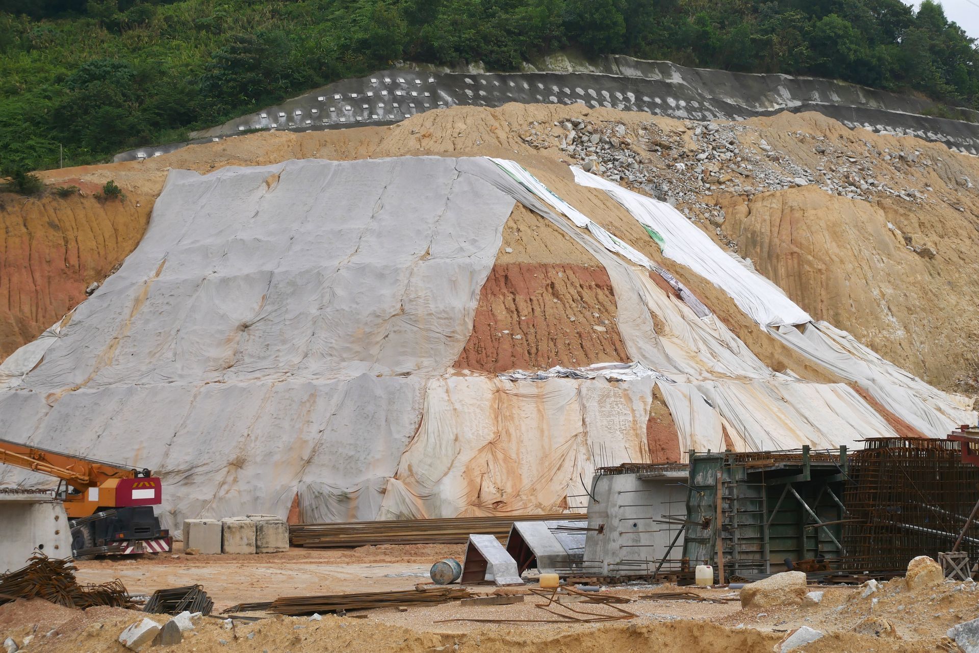 Construction site: Earth and rock hillside covered with white protective material, with a crane in the foreground.