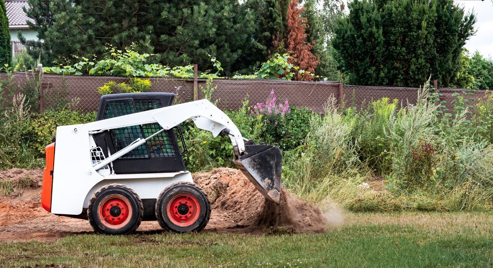 White Bobcat skid-steer loader scooping up dirt in a grassy area with a fence and trees in the background.