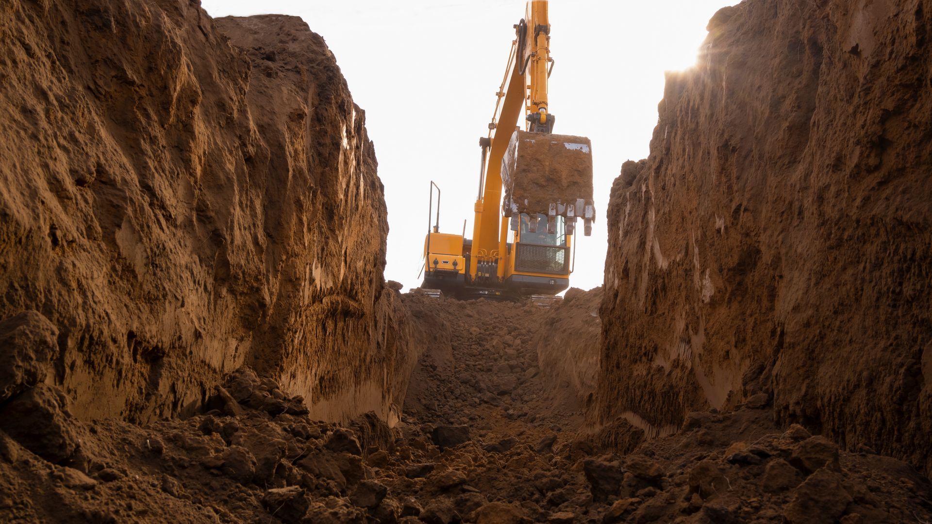 Yellow excavator digging in a deep trench, bright sky in the background.