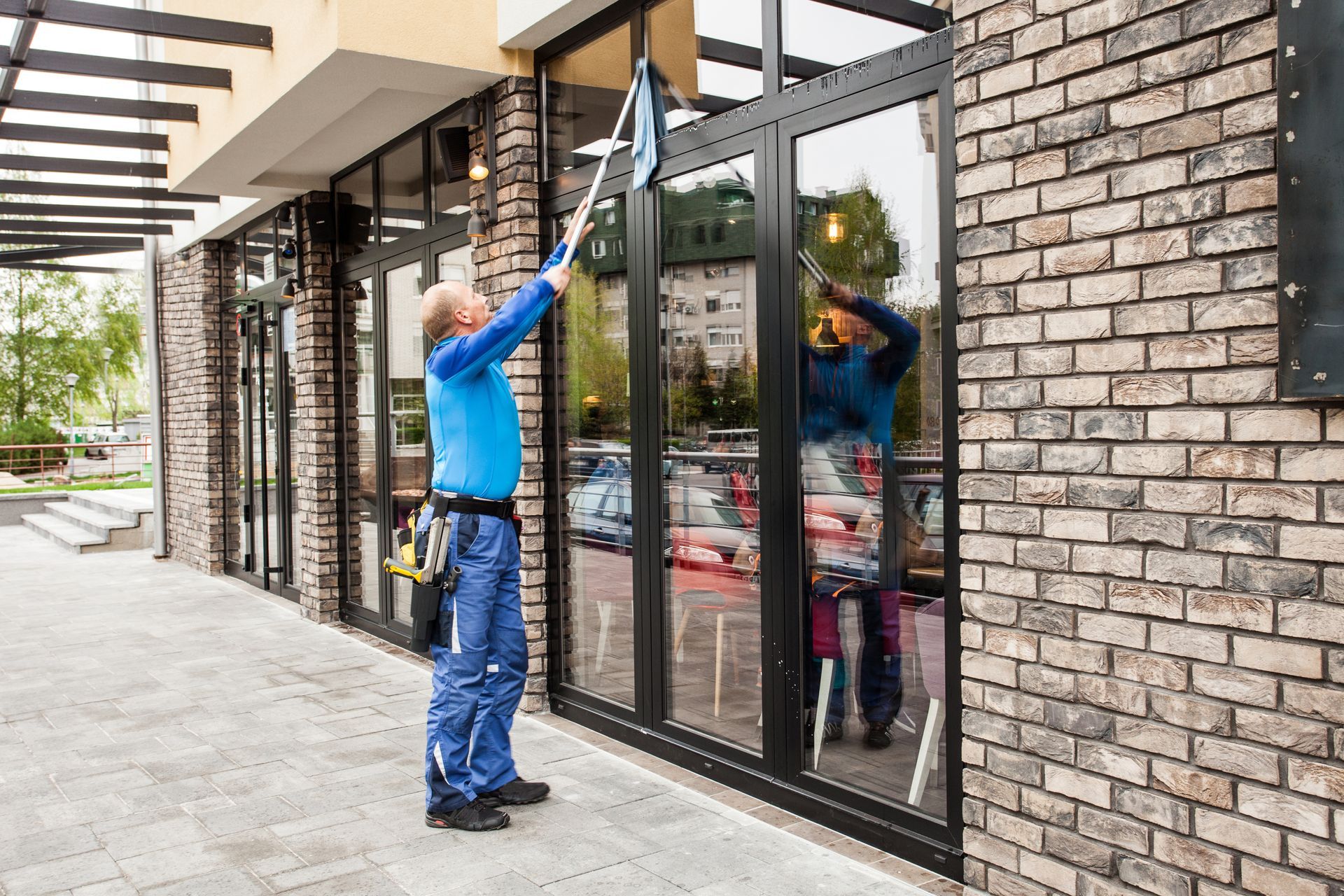 Worker on a ladder cleaning large floor-to-ceiling windows inside a bright building.
