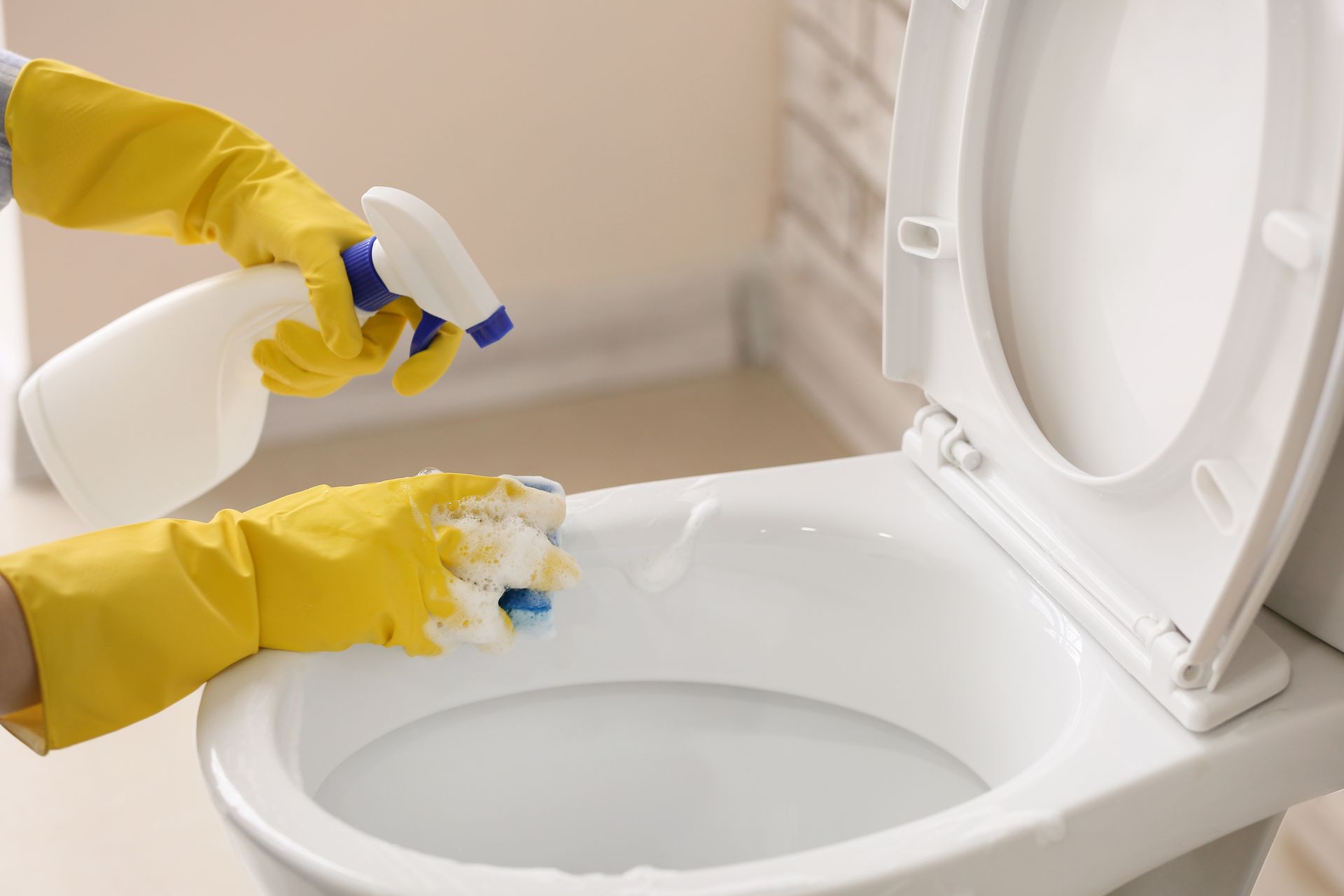 Gloved hands spraying and scrubbing a white toilet with cleaner in a bathroom