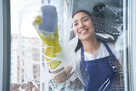 Woman in blue apron cleaning a window with a sponge, smiling amid soap suds.