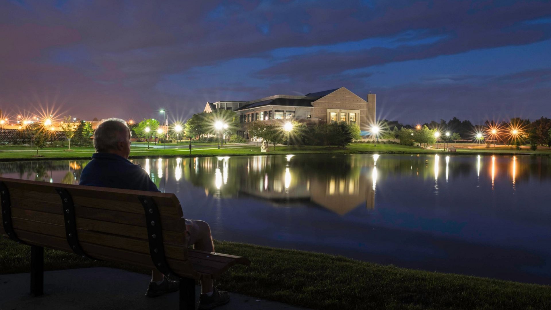 Person sitting on a bench by a lake at dusk, with building lights reflecting on the water