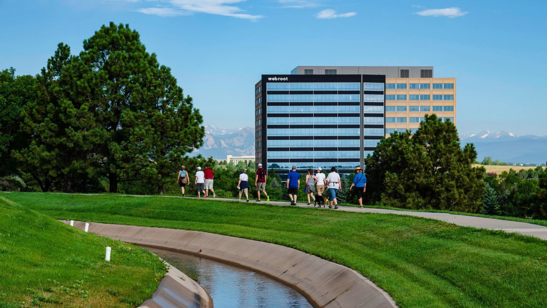 People walking on a grassy path near a modern office building under a blue sky.