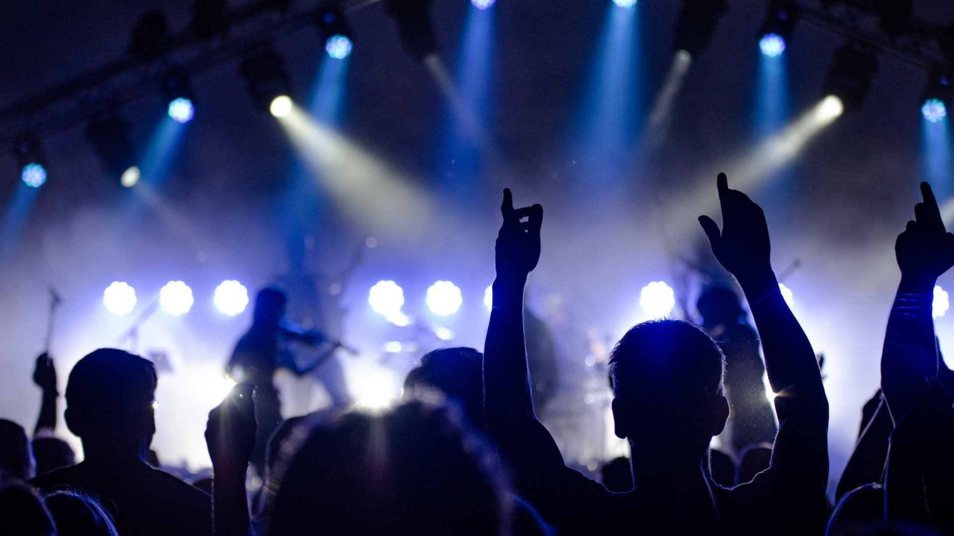 Crowd silhouetted at a concert, arms raised under blue and white stage lights.