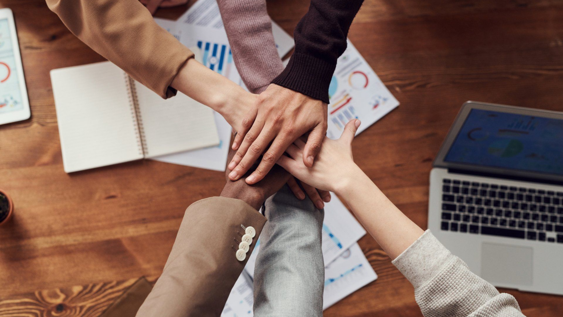 Hands stacked in a team huddle over a wooden table with papers and a laptop nearby