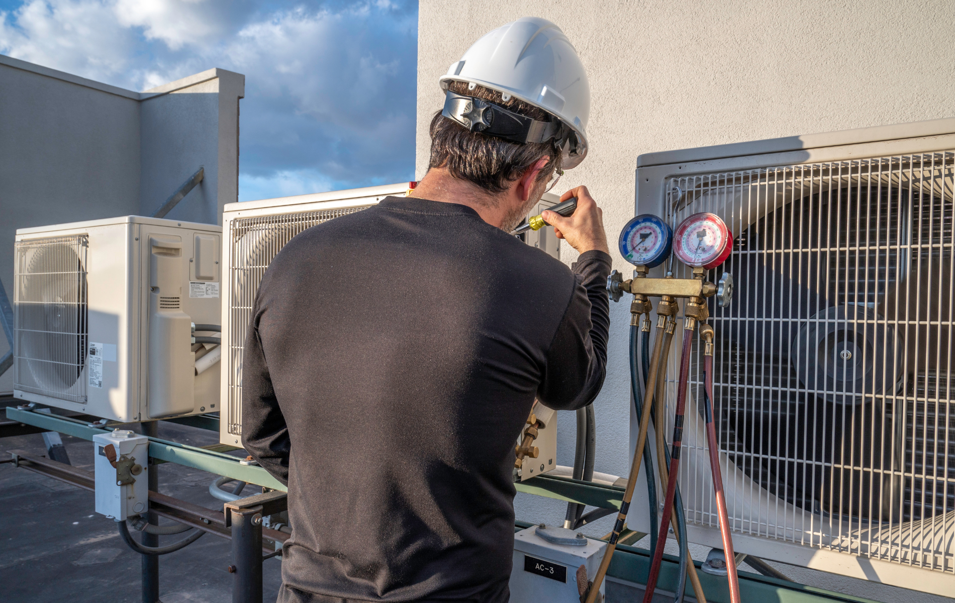 Technician in hard hat inspecting rooftop HVAC units with gauges and tools under a cloudy sky