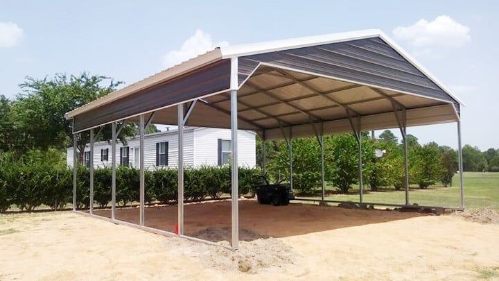 Open-sided metal carport beside a small white building on a sandy lot, with trees in the background