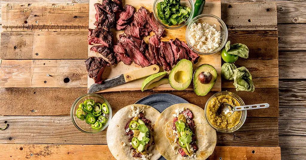 A wooden table topped with tacos , guacamole , meat and vegetables.