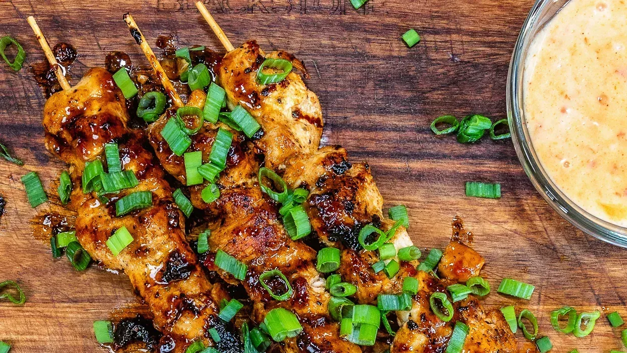 A wooden cutting board topped with chicken skewers and green onions next to a bowl of dipping sauce.