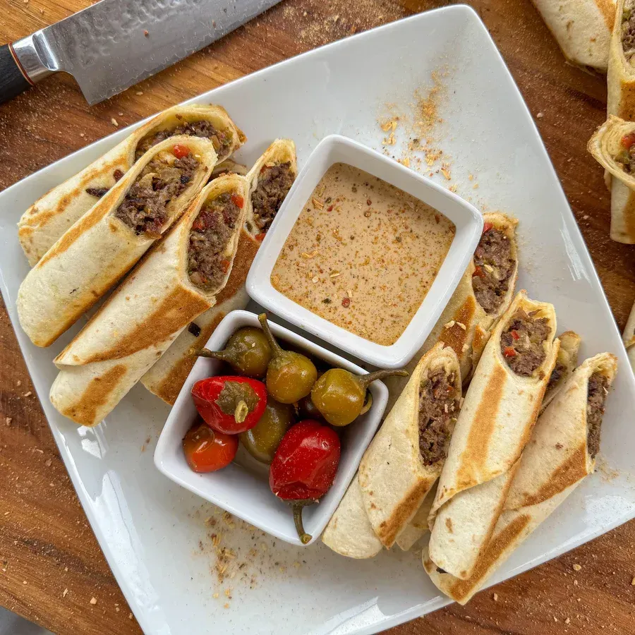 Plate of filled tortilla rolls with dipping sauce and pickled peppers.