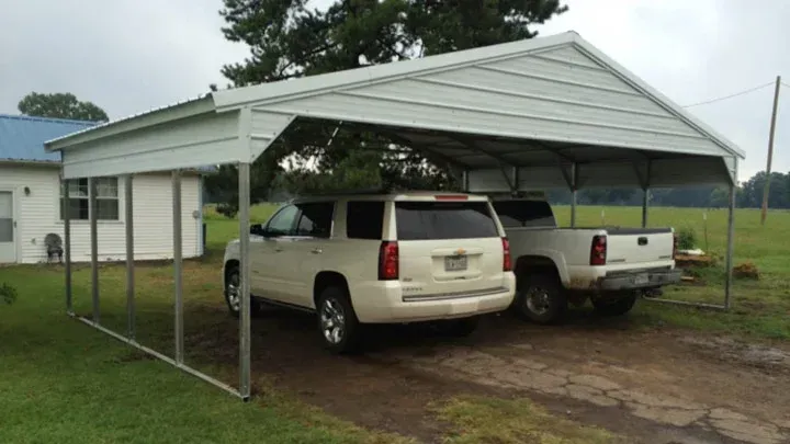 Two pickup trucks parked under a metal carport beside a small house on a muddy lot