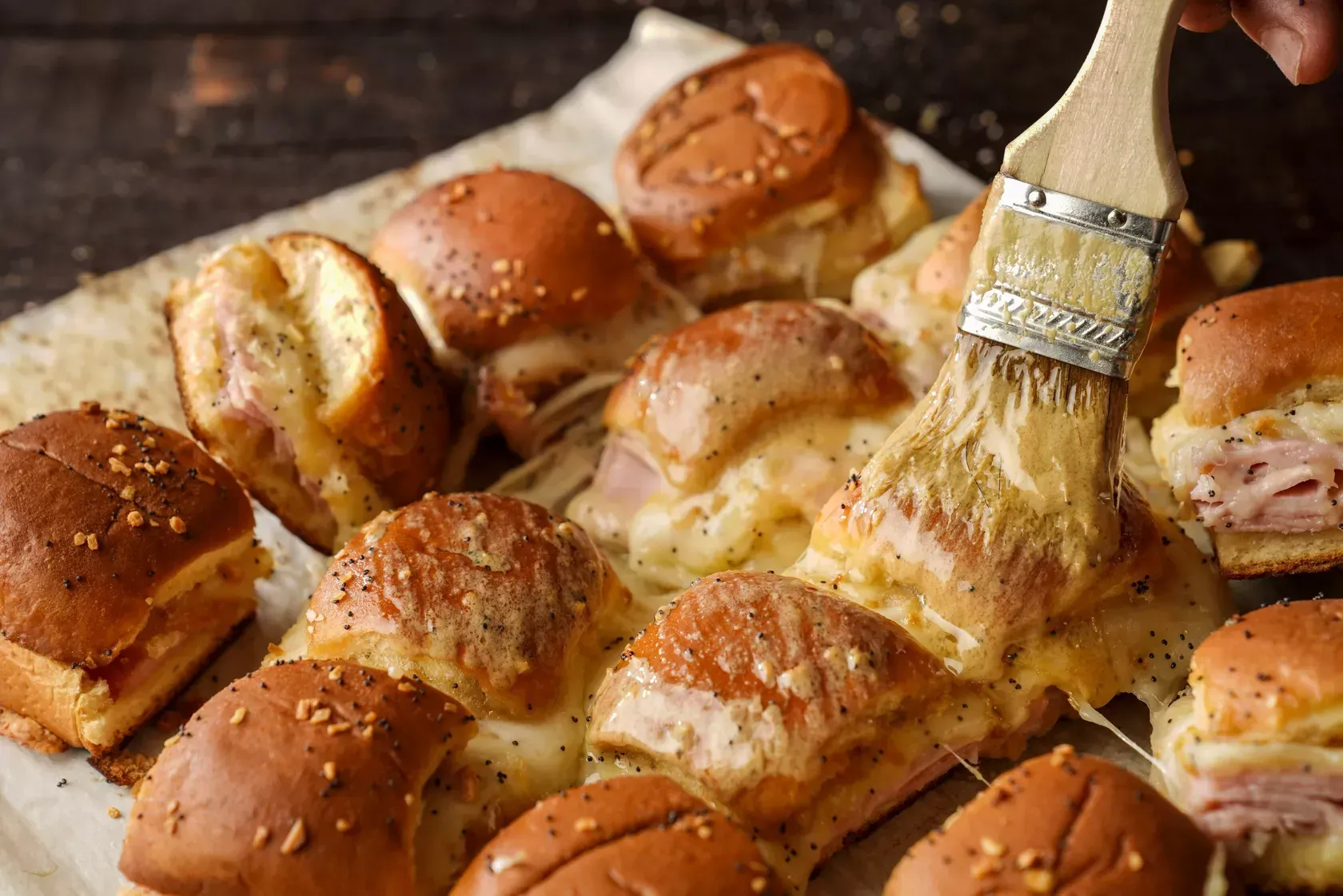 Baked sliders being brushed with a buttery sauce, melting cheese visible, on parchment paper.