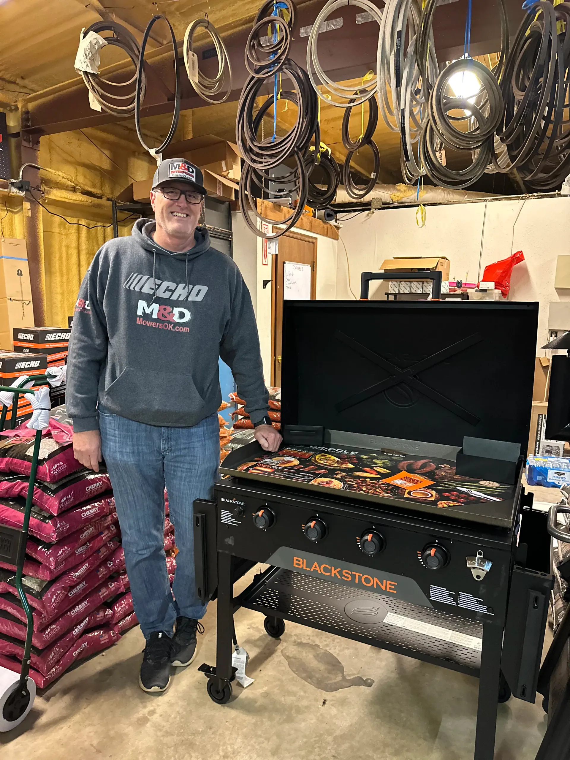 Man standing beside a black grill in a garage, with coiled hoses and tools hanging overhead.