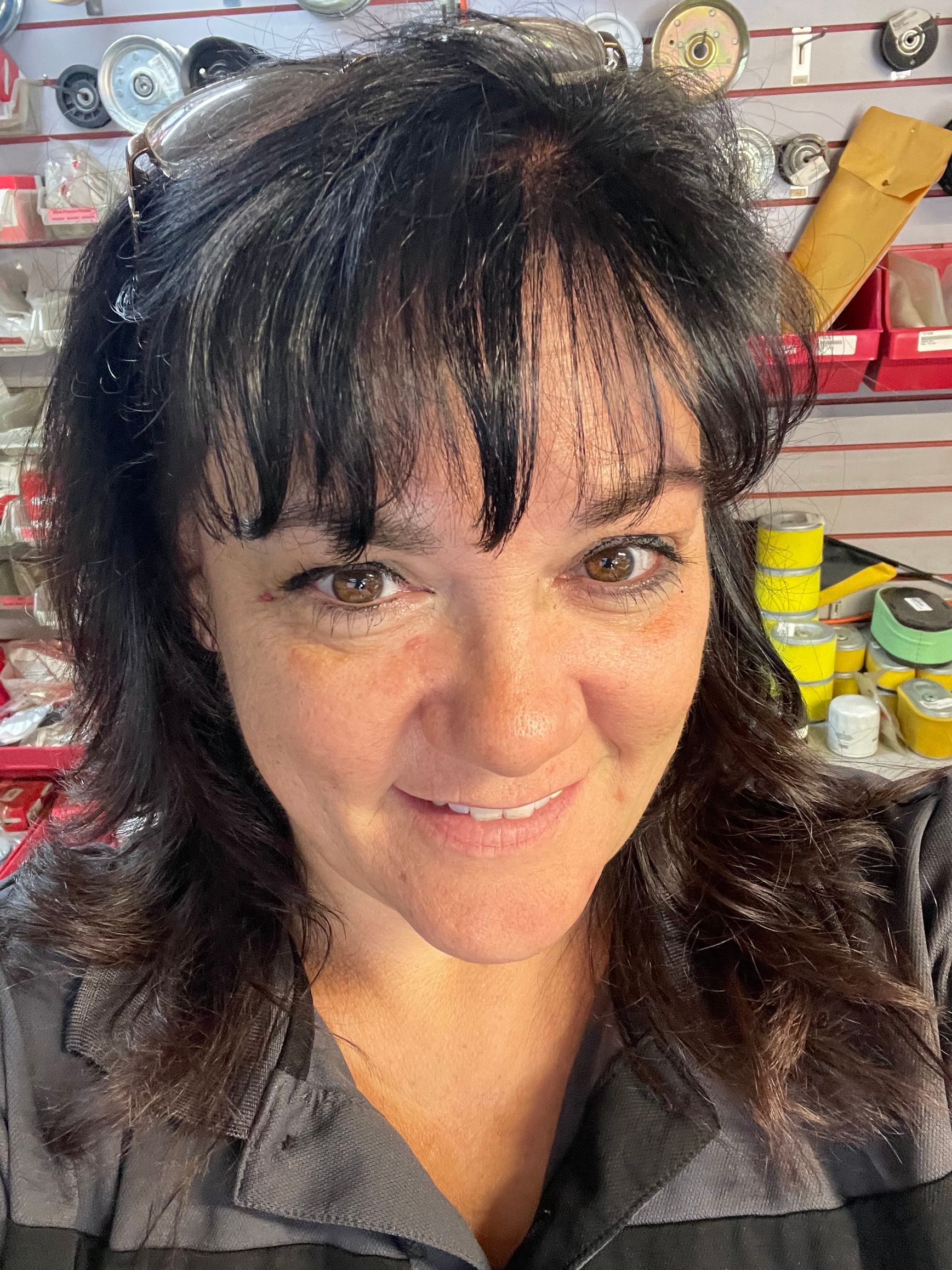 Smiling person with dark hair in a workshop, with shelves of tools and supplies behind them.