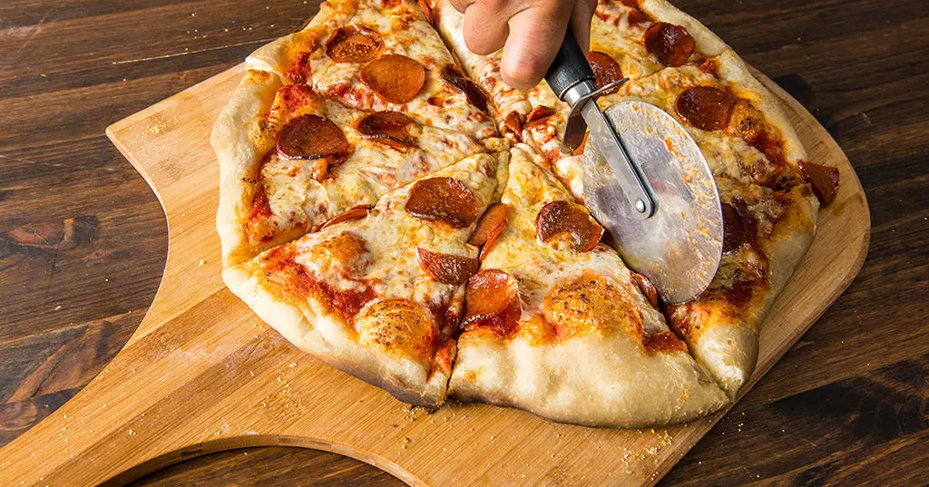 A person is cutting a pepperoni pizza on a wooden cutting board.