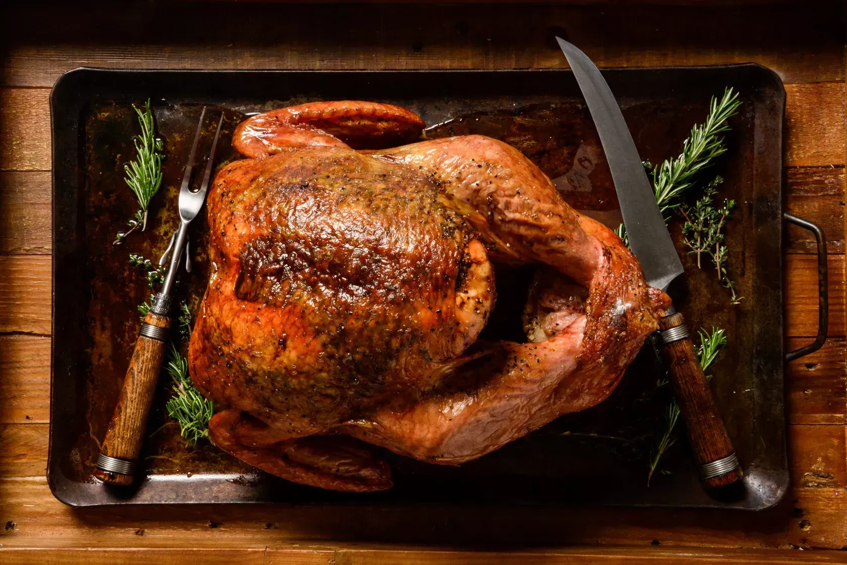 Roasted turkey on a tray with knife, fork, and rosemary sprigs on a wooden table.