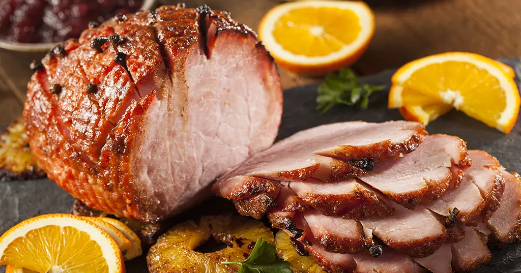 A ham is sitting on a cutting board next to oranges and pineapple slices.