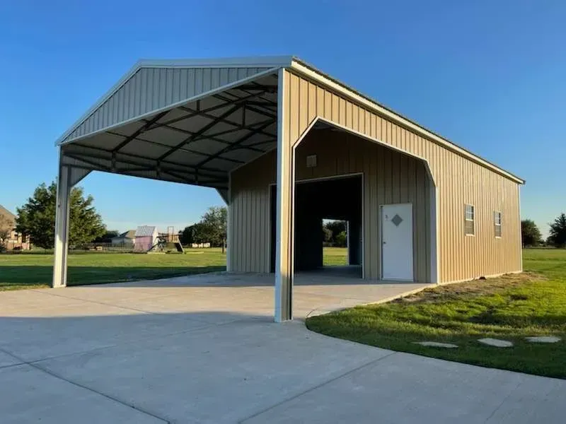 Open-sided tan metal pavilion with a gray roof beside a concrete driveway on a grassy lot