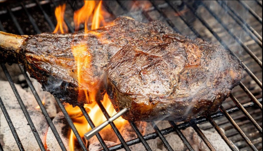 A large steak is cooking on a grill with flames.