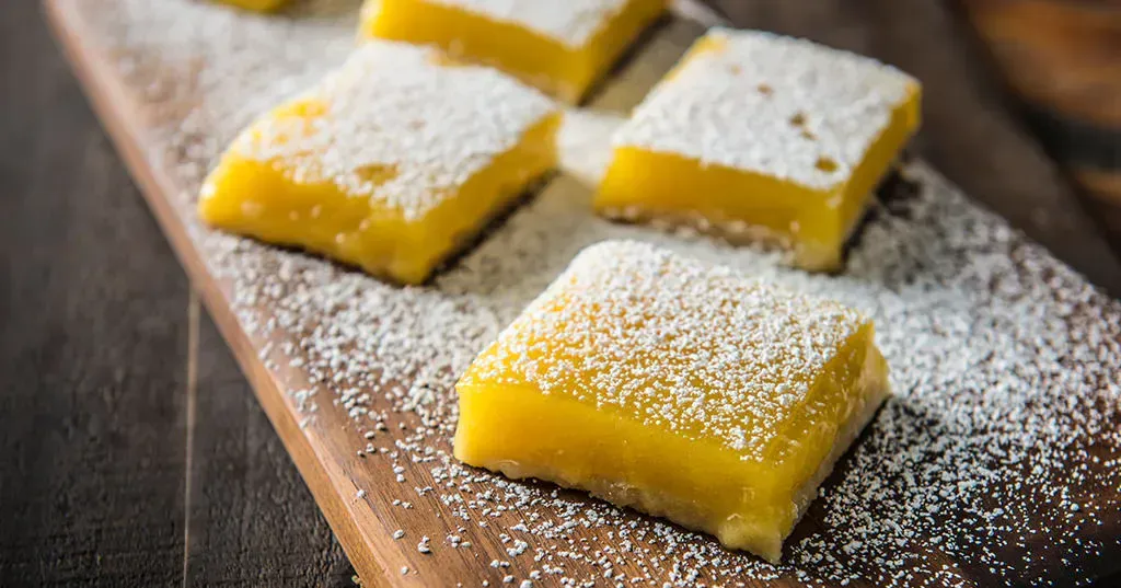A wooden cutting board topped with lemon bars covered in powdered sugar.