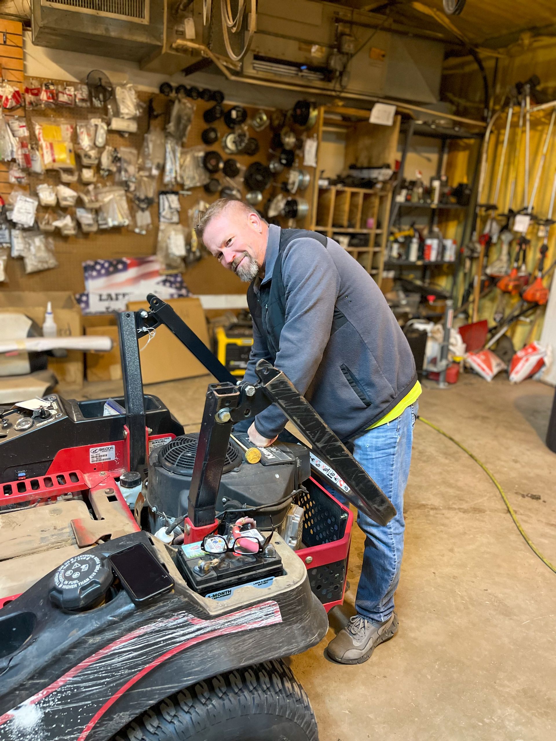 Person working on a red and black lawn mower in a cluttered garage workshop