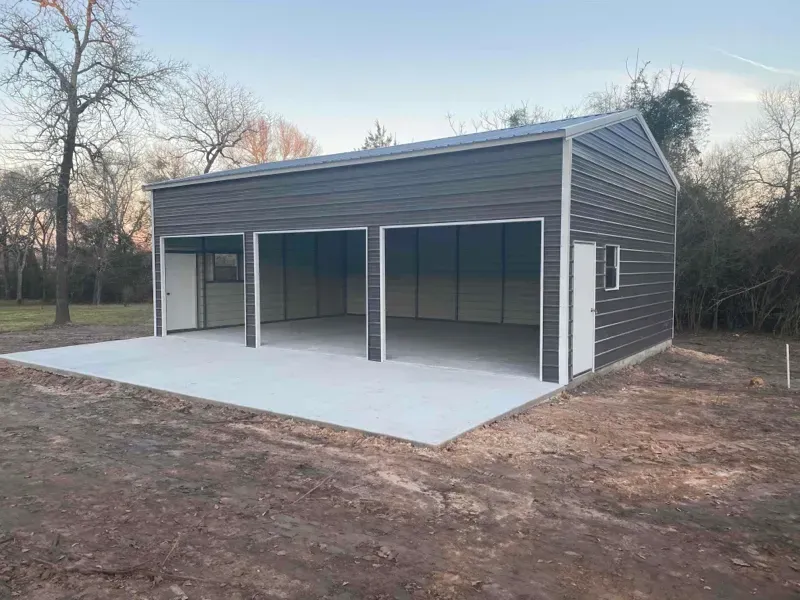 Gray three-bay metal garage with white concrete pad in a wooded yard