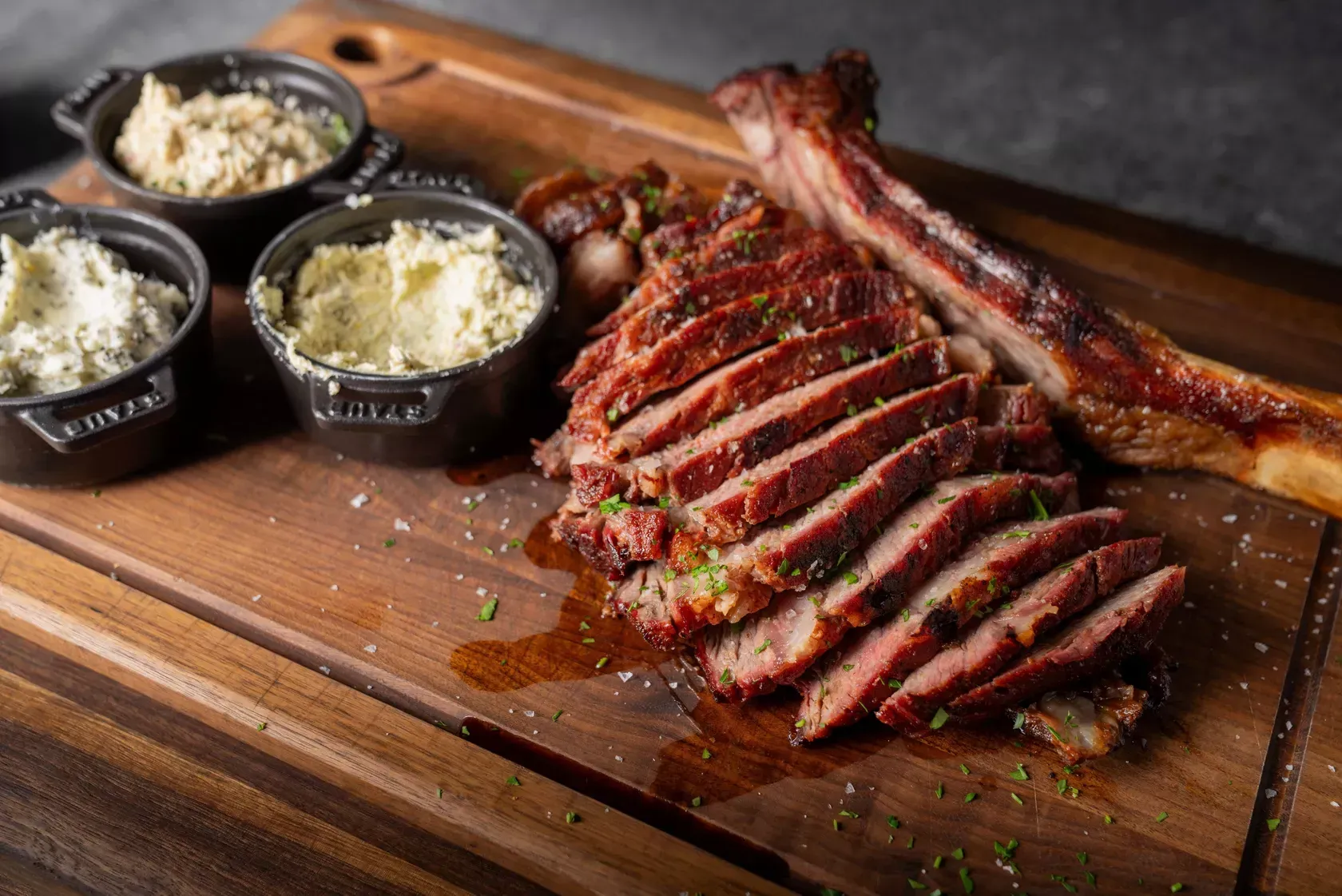 Sliced grilled steak with bone, served with three small bowls of sauces on a wooden board.