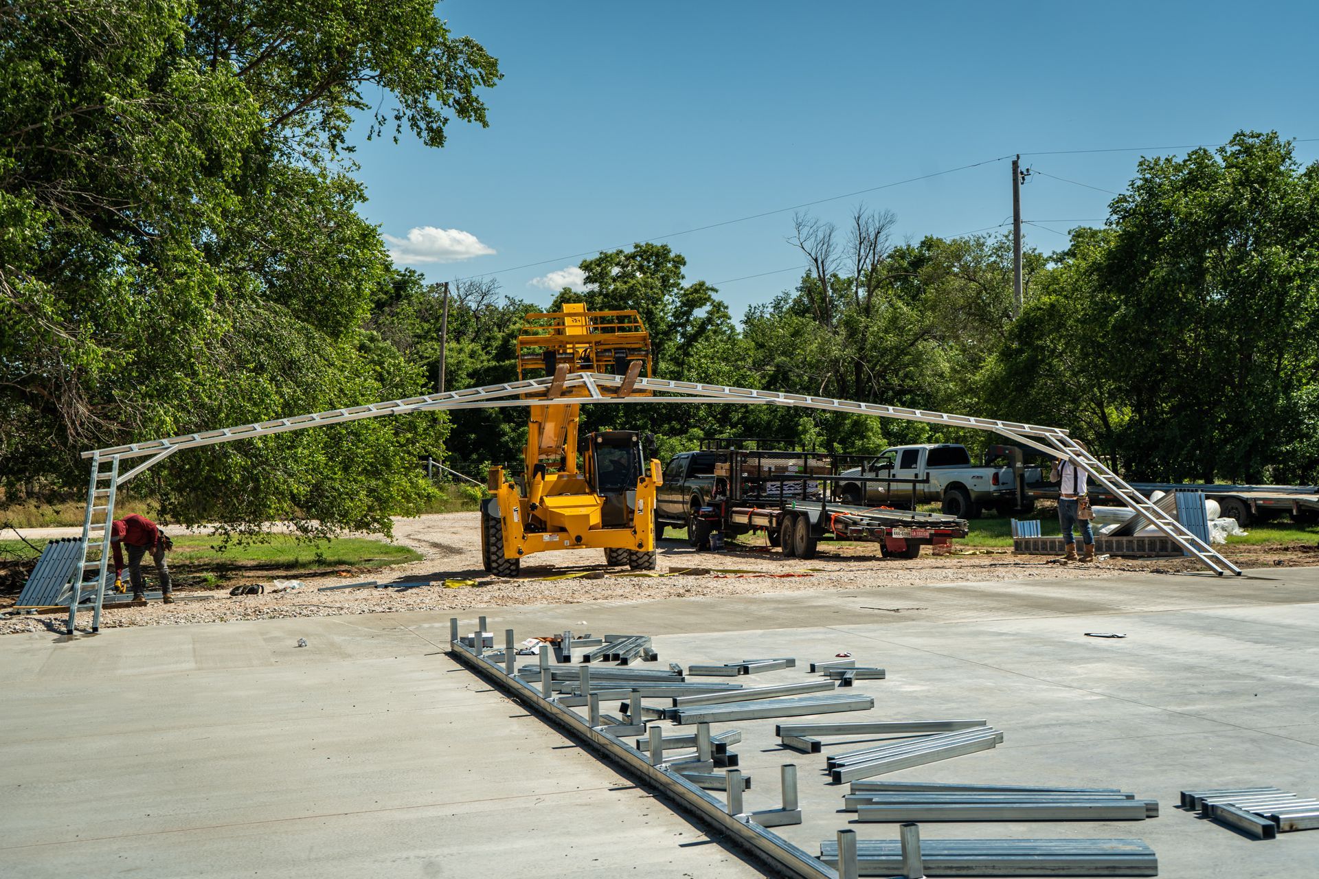 Construction site with a crane lifting a curved metal frame on a concrete slab surrounded by trees