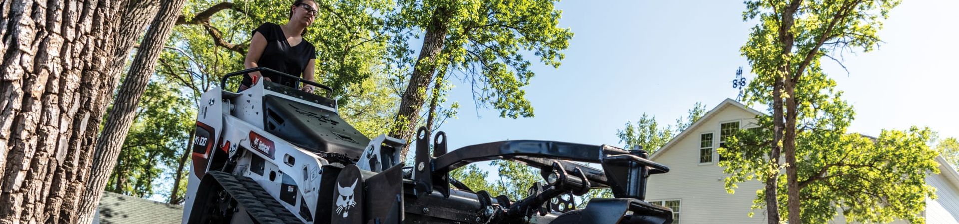 a man is standing on top of a mini track loader next to a tree .