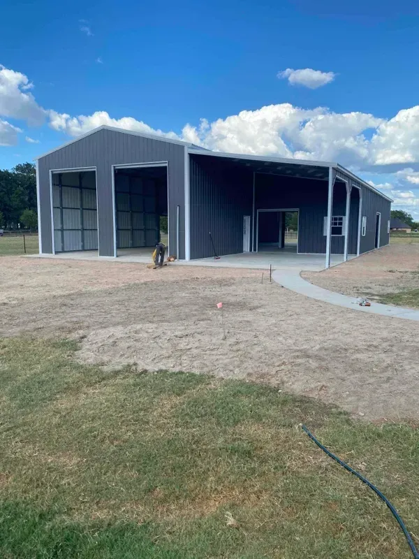 Blue metal barn-like building with open bays, on a grassy dirt lot under a partly cloudy sky.