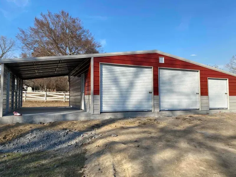 Red metal barn with three white garage doors and an open side shed on a dirt lot under a blue sky