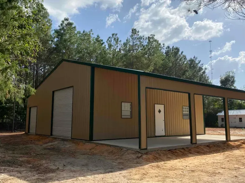 Tan metal warehouse with roll-up doors and covered porch on a dirt lot beside trees