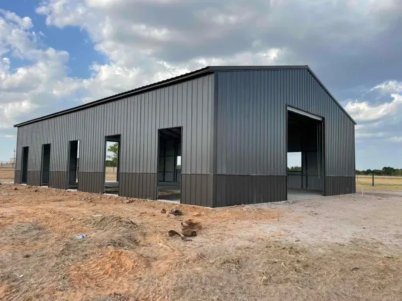 Dark gray metal building under construction on a dirt lot, with open window and door frames
