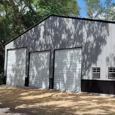 Gray metal storage building with three roll-up doors and small windows, surrounded by trees.