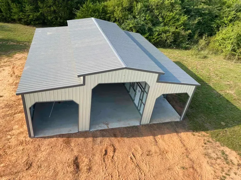 Small gray metal barn with three open bays, set on bare dirt beside green trees.