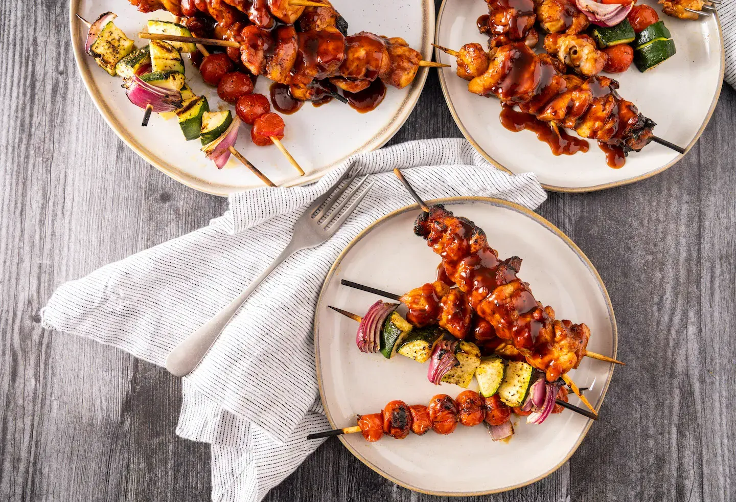 Three plates of skewers of meat and vegetables on a wooden table.