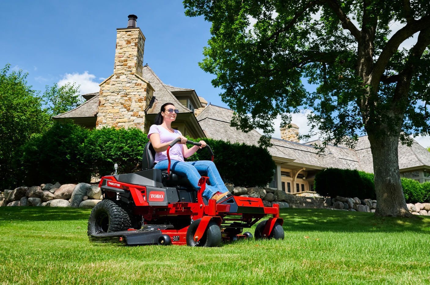 Person riding a red riding lawn mower on a green lawn in front of a stone house
