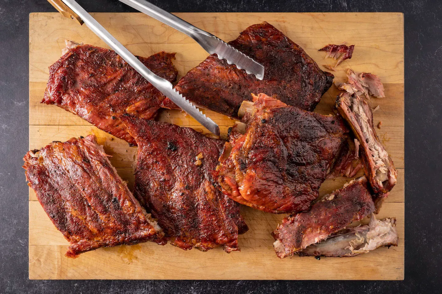 A wooden cutting board topped with ribs and tongs.