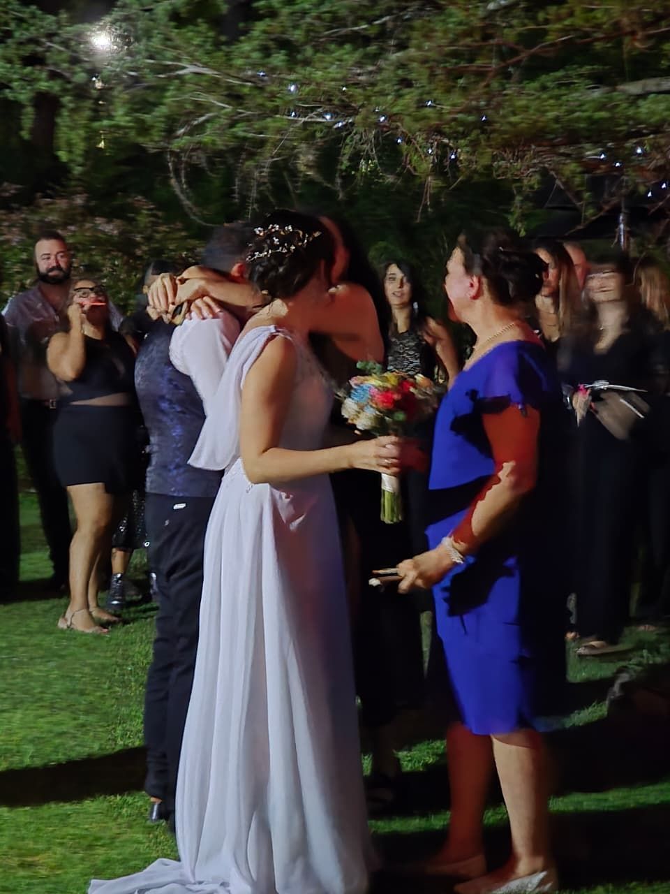 Invitados a una boda se abrazan al aire libre al atardecer, con una novia vestida de blanco.