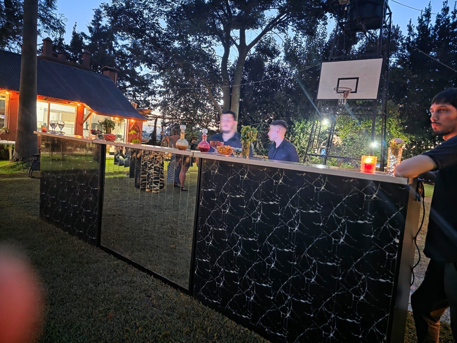 Reunión nocturna al aire libre con gente detrás de una barra de mármol negro iluminada y una canasta de baloncesto cerca.