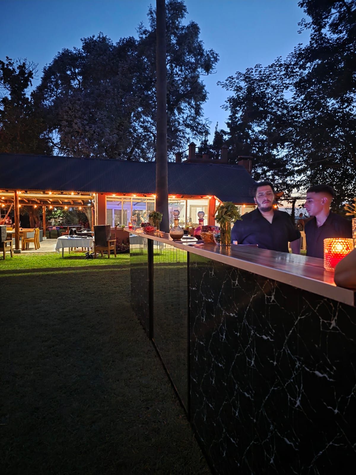 Un bar al aire libre al atardecer con dos personas charlando junto a una barra iluminada.